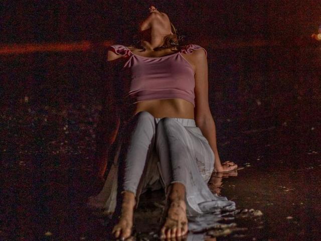 NaturalMariaHun's head is tilted back, mouth slightly open, in a pink crop top, showing her small tits, and blue jeans, sitting with dirty feet in dark water.