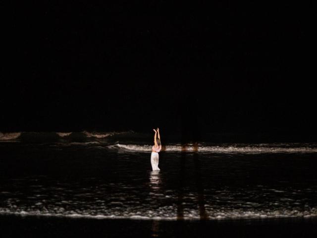 Night photo of NaturalMariaHun standing in water, arms raised, wearing white dress, small tits visible, dark background, reflection on water, slight wave, isolated, centered, serene.
