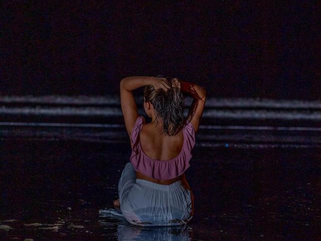 NaturalMariaHun with wet hair, arms raised, wearing a pink top and white pants, kneeling in dark water, back to the camera, reflecting on the surface.