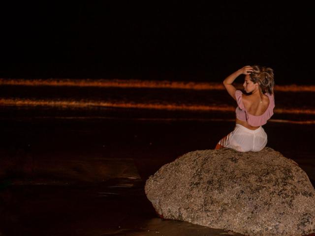 NaturalMariaHun está sentada sobre una roca por la noche, vestida con una blusa rosa y una falda blanca, con la mano en el pelo, de espaldas a la cámara, mostrando su trasero..