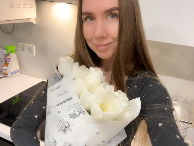 SannyManny with long brown hair, green eyes, and a slight smile, holding a bouquet of white tulips, wearing a black star-patterned top, in a kitchen.
