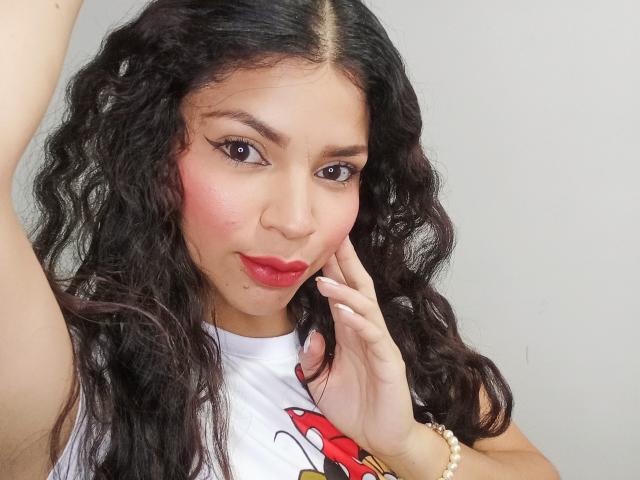 EstrellaHotG with curly black hair, pink blushed cheeks, red lipstick, and white graphic tee, touching her face, wearing a gold bracelet, and looking at the camera.