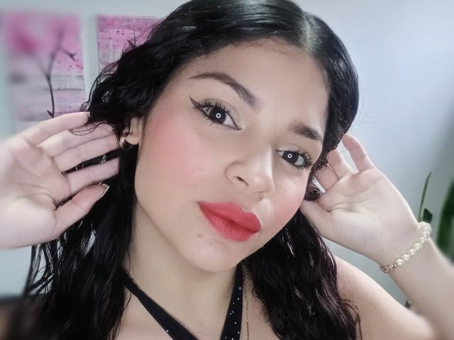 EstrellaHotG has black hair, red lipstick, and is touching her ears with hands. She's wearing a black halter top and a pearl bracelet, with a cute, sexy look.