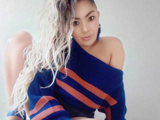 KarenCurlyHair with long blonde hair, wearing a blue off-shoulder sweater with red stripes, sitting, showing bare shoulders and black shorts, smiling slightly.
