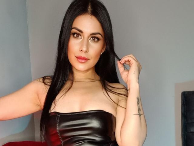 MaddySmit, con i suoi lunghi capelli neri, indossa un top nero in pelle senza spalline, ha un tatuaggio sul braccio destro e sta toccando i capelli con un leggero sorriso..