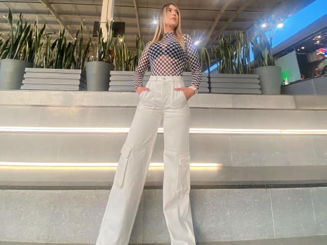 AlinaSmiith stands with hands in white cargo pants, wearing a checkered top, under a ceiling with lights, in front of large potted plants, looking confident.