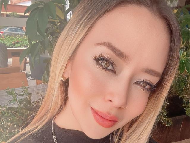 AlinaSmiith has blonde hair, light skin, brown eyes with long lashes, and red lipstick. She's smiling with a nose ring, wearing a black top and gold earrings.