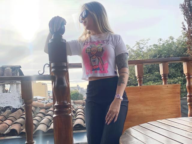 AlinaSmiith with long blonde hair, wearing sunglasses, white crop top with pink cartoon, black high-waisted pants, standing on wooden balcony, touching wooden post, tattoo on right arm.