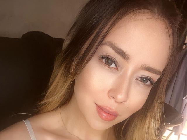 AlinaSmiith has light brown skin, green eyes, long eyelashes, and glossy pink lips. She's looking at the camera with a slight smile, and her brown hair is loose.