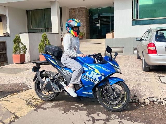 AlinaSmiith in a blue Suzuki sport bike, wearing a colorful helmet, gray outfit, and white sneakers, parked next to a silver car, in a sunny urban setting.