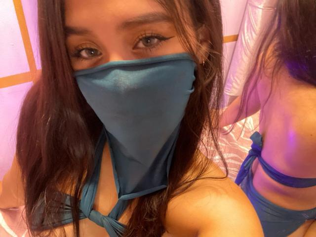 EmyXCute mit langen braunen Haaren, die einen blauen Maske und ein passendes blaues Bikini trägt, zeigt ihre Brüste und ihren Hintern in einem Spiegel-Selfie. Ihre Augen sind mit Eyeliner betont..