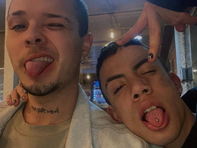 AronJames, con un anello al naso e un tatuaggio, che mostra la lingua. Un altro AronJames, anch'esso con un anello al naso, ha la lingua fuori e viene toccato da un dito..