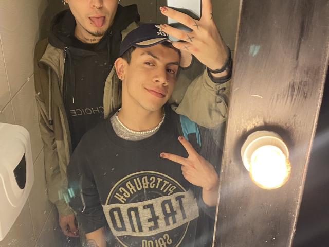 AronJames en un selfie frente al espejo con otro chico, con la lengua afuera. AronJames, con una camisa negra que dice "Quartic", un collar de perlas y un gorro, haciendo el gesto de la paz. Un chico vestido está detrás..