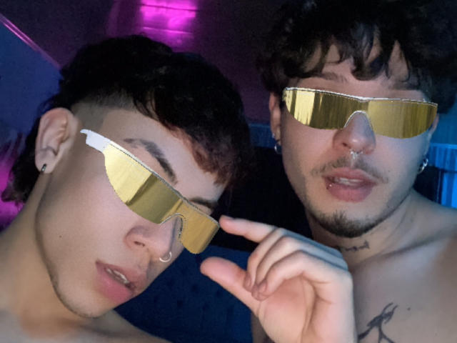 AronJames and AronJames wearing shiny gold aviator sunglasses, both with piercings and tattoos, dark curly hair, and lip piercings, in a dimly lit, purple-lit room.