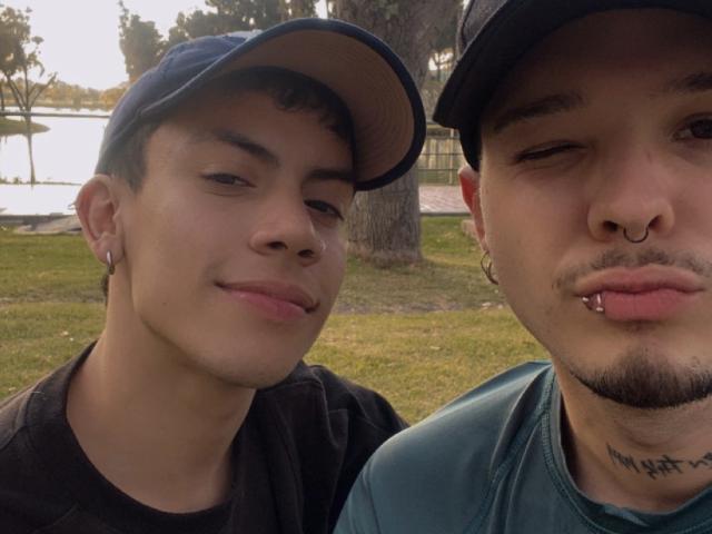 AronJames, con cabello negro corto, gorra, camisa negra y perforaciones en las orejas, está de pie junto a AronJames, que tiene bigote, aro en la nariz, piercing en el labio y un tatuaje, y que lleva una camisa verde..