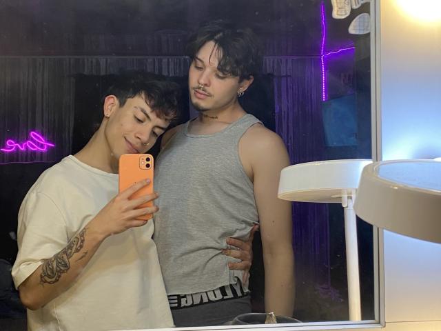AronJames with short dark hair, white tee, tattoo on left arm, holding phone, smiling, leaning on AronJames in grey tank, choker, black pants, standing in front of mirror.