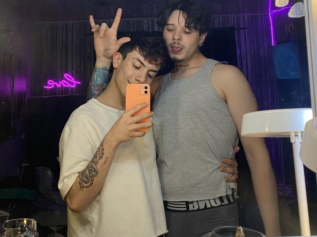AronJames with tattooed arm, white tee, and AronJames with curly hair, nose ring, grey tank, and PUMA underwear, both making silly faces in a mirror selfie.