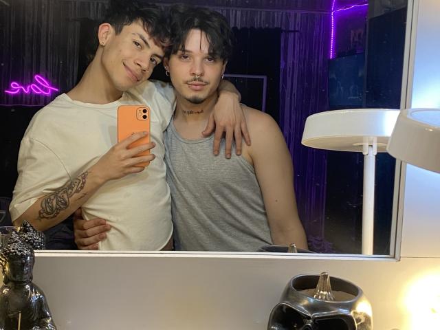AronJames with tattooed arm and white tee, holding phone, stands next to AronJames in gray tank, arm around. Mirror selfie, purple neon, white lamp, black statue, and metal holder.