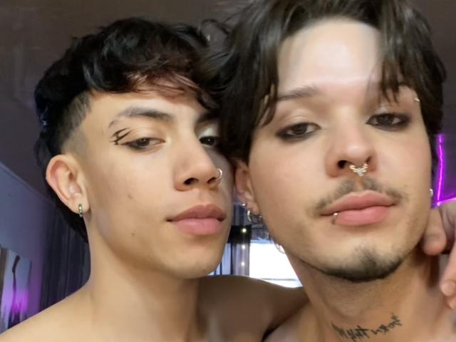 AronJames with short black hair and nose ring, standing next to AronJames with brown hair, nose ring, and lip piercing, both showing off their piercings and tattoos.
