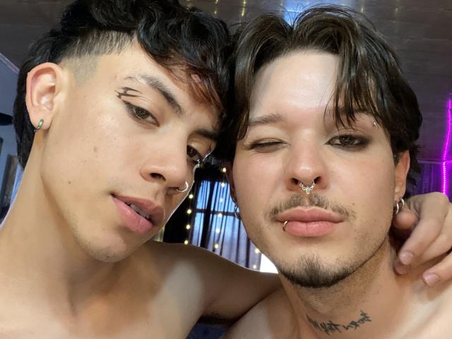 AronJames with short black hair and nose ring, next to AronJames with brown hair, nose ring, and mustache, both shirtless, looking at camera, arms around each other.