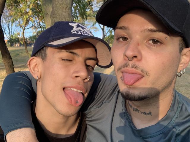 Two dudes with black caps, nose piercings, and tongue piercings, sticking out tongues, arms around each other, one with tattoos on neck, in a sunny park.
