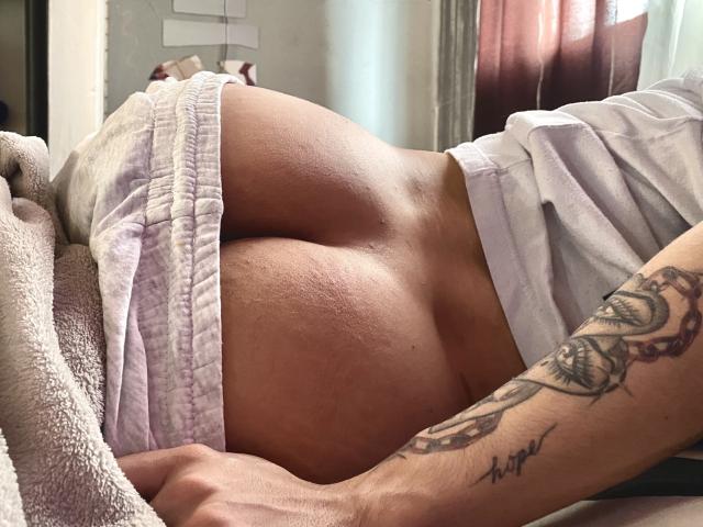 AronJames is lying down with their bare ass exposed, wearing white shorts pulled to the side, showing a tattooed arm and a soft, textured blanket.