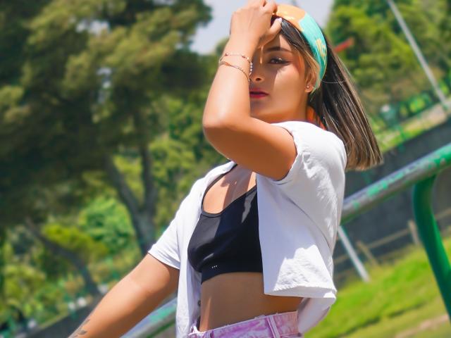 CloeesSuarez with a tan skin tone, wearing a white shirt, black sports bra, and pink pants, adjusting a green headband, showing her tits and looking sexy.