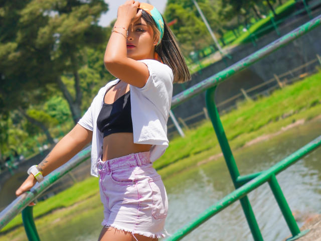 CloeesSuarez, met een bruine huidskleur, een zwarte top, een witte jas, roze denim shorts en een groene hoofdband, die haar haar aanpast, staat bij een groene reling, met een zonnig park op de achtergrond..