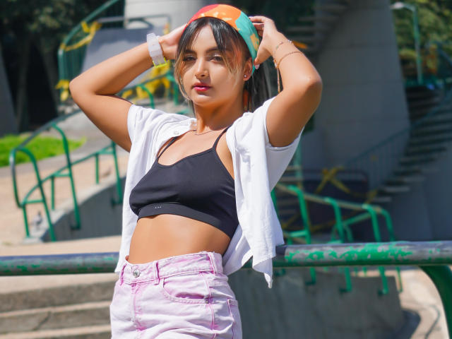CloeesSuarez with small tits, wearing a black sports bra, white open shirt, pink high-waisted jeans, and a rainbow cap, standing with arms up in a sunny park.