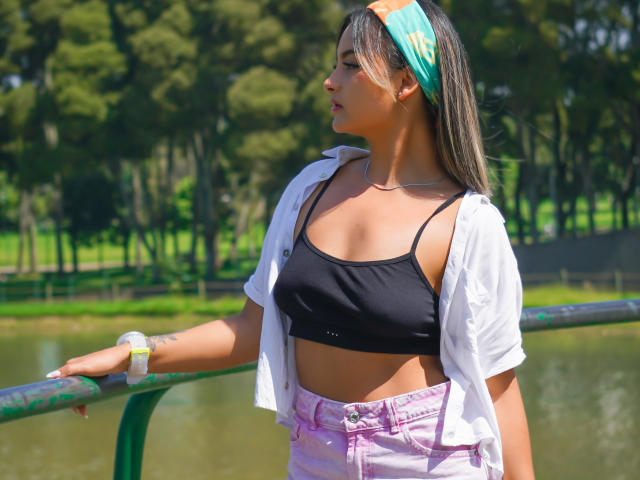 CloeesSuarez with tanned skin, small black bra, white open shirt, pink pants, green headband, leaning on green railing, tattoo on left wrist, standing by a pond.