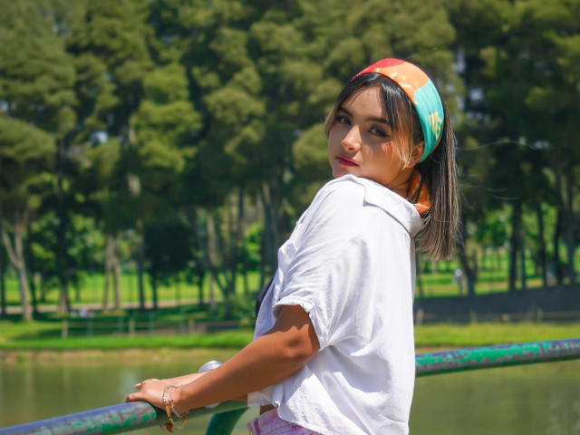 CloeesSuarez with short black hair, white shirt, colorful headband, looking back over shoulder, standing by green railing, wearing bracelet, by sunny park with trees.