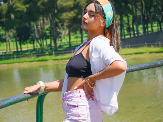 CloeesSuarez with titts in a black bra, white shirt, and pink pants, leaning on a green railing by a pond, wearing a green headscarf and bracelets, looking up.