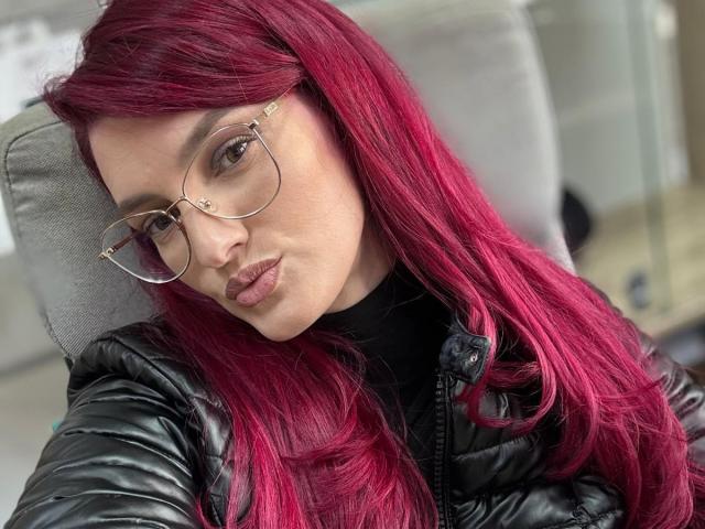 Rosalyn with pink hair, wearing glasses and a black puffy jacket, pouting her lips, showing off her tits in the selfie. She's looking directly at the camera.