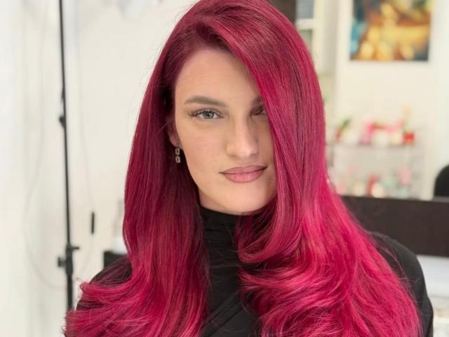 Rosalyn, com o cabelo rosa vibrante, piscando, vestindo um suéter preto de gola alta, sorrindo levemente, exibindo seus olhos verdes e maquiagem discreta. O cabelo de Rosalyn é longo e brilhante..