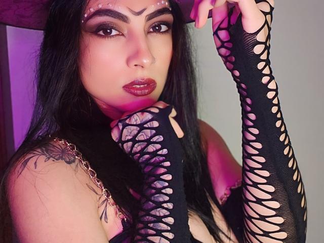 HhMeghanOlim with long black hair, dark makeup, and red lipstick, wearing black lattice gloves and a black top, showing a tattoo on her shoulder and holding her chin.