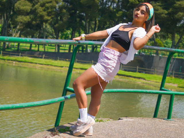 CloeesSuarez met een bruine huid, een zwarte sportbeha, een wit shirt, roze shorts, witte sokken en sneakers, leunt tegen een groene reling, draagt een kleurrijke hoofdband, bij een zonnig parkmeer..