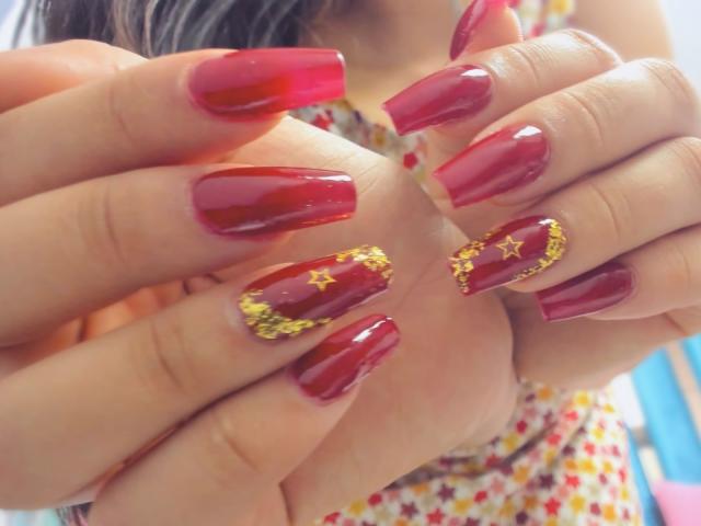 Genesiss' hands with shiny red nails, some adorned with gold star patterns. Her fingers are slightly curled, showcasing the detailed nail art. Her skin is light, and her nails are glossy.