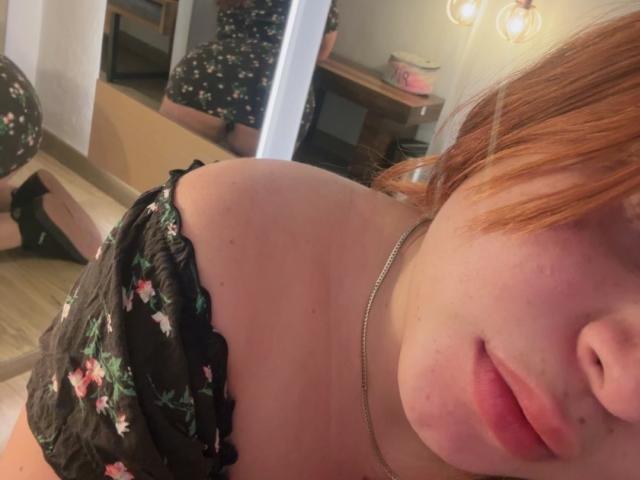 RoseDijon with red hair, wearing a black floral off-shoulder top, showing her bare shoulder and upper chest, a reflection of her ass in a mirror, and a necklace.