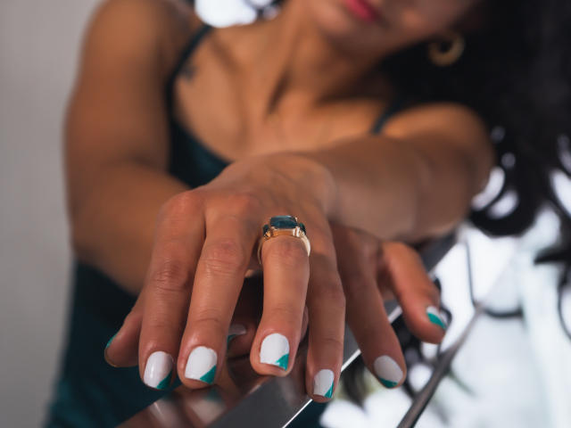 EvelinLuna's hand with a gold ring, white and teal nail polish, and green dress, reaching forward, showing off her manicured nails and shiny ring. Blurred face in background.