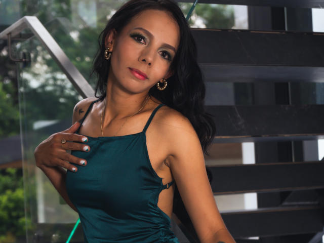 EvelinLuna with dark hair, green dress, gold earrings, hand on chest, looking at camera, medium tits, slight smile, pink lipstick, sunlight, black stairs, glass, greenery outside.