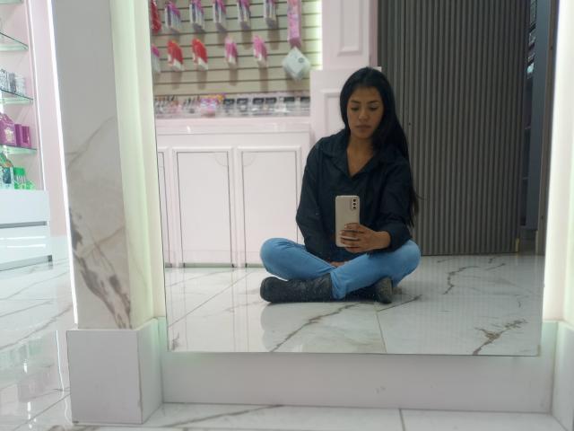 ArianithaXox está sentada de pernas cruzadas em um chão de mármore branco, vestindo uma jaqueta preta e jeans azuis, tirando uma selfie no espelho com um smartphone. Suas nádegas estão parcialmente visíveis..