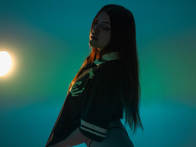 MaylinSanchez with brown hair, wearing a green Vandepf shirt, flexing arms, nose ring, and dark makeup, standing against a blue gradient background.