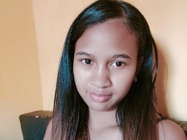 LoderFist has dark brown straight hair, medium brown skin, small earrings, and a subtle smile, showing brown eyes and pinkish lips, with a slight sheen on her face.