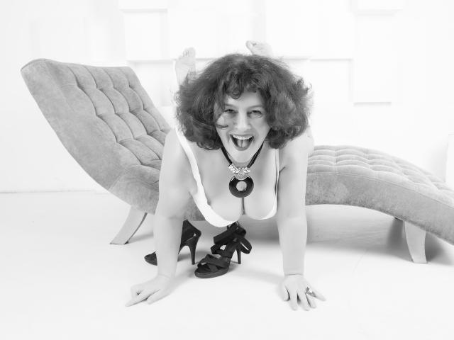 BerryChic with curly hair, tongue out, leaning on a tufted chaise, wearing a white top, necklace, and high heels, her big tits and ass slightly visible. Black-and-white photo.