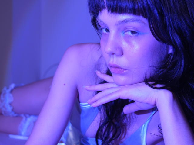 GodessSam with blue-tinted skin, long black hair, bangs, wearing a silky blue top, resting her chin on her hand, looking off to the side, with a serious expression.
