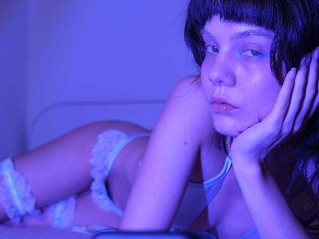 GodessSam with blue-lit skin, lying on her side in white lace panties and garter, hand under chin, looking at camera, dark bangs, glossy lips, soft expression.