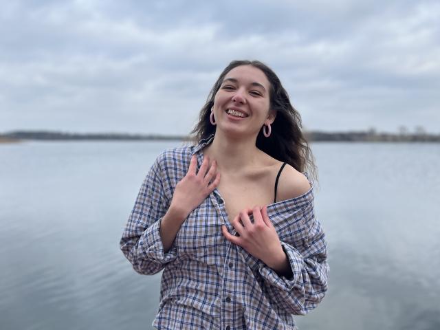 KissMegan with long brown hair, smiling, wearing a plaid shirt, hands on chest, showing black bra strap, pink earrings, standing by a calm water body, overcast sky.