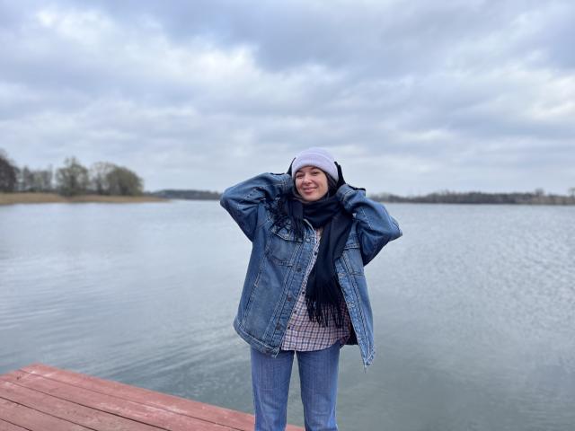 KissMegan debout sur une jetée en bois, portant une veste en jean bleu, un bonnet blanc, une écharpe noire, une chemise à carreaux et un jean bleu, les mains dans les poches de la veste, ciel nuageux, eau calme..