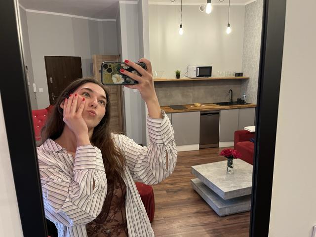 KissMegan, avec de longs cheveux bruns, une chemise rayée et des ongles rouges, tenant un téléphone, faisant une moue, selfie devant un miroir, sol en bois, cuisine moderne, chaise rouge et petite table avec des fleurs..
