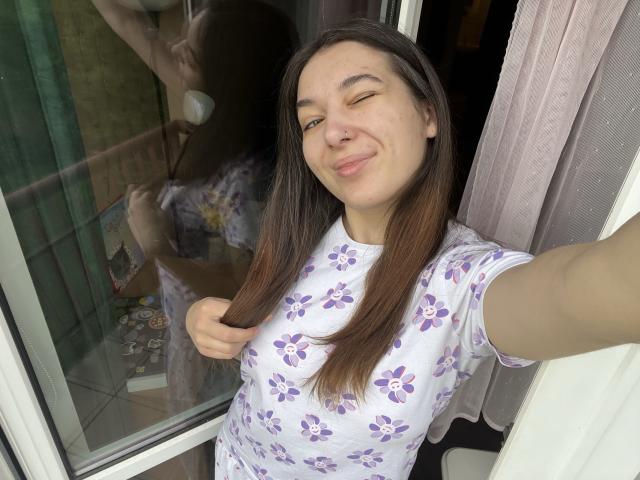 KissMegan, avec de longs cheveux bruns, portant une chemise blanche à motifs floraux, souriant les yeux fermés, tenant ses cheveux pour un selfie, montrant un anneau de nez mignon et un visage détendu..